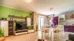 Un salon moderne doté d’un téléviseur à écran plat, d’un coin repas avec des chaises blanches et d’une cuisine avec des meubles violets.