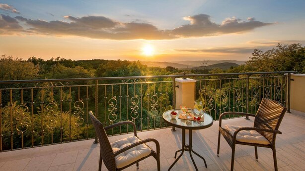 En balkon med fletstole og et bord dækket til to, med udsigt over et naturskønt landskab i solnedgang.