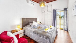 Un dormitorio con techo de madera, una cama con ropa de cama estampada, una mesita de noche con lámpara, una silla roja y un ventanal corredizo que da a un área exterior.