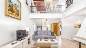 Un amplio salón con sofá, mesa de centro y televisor. Una escalera conduce a un balcón con barandilla.