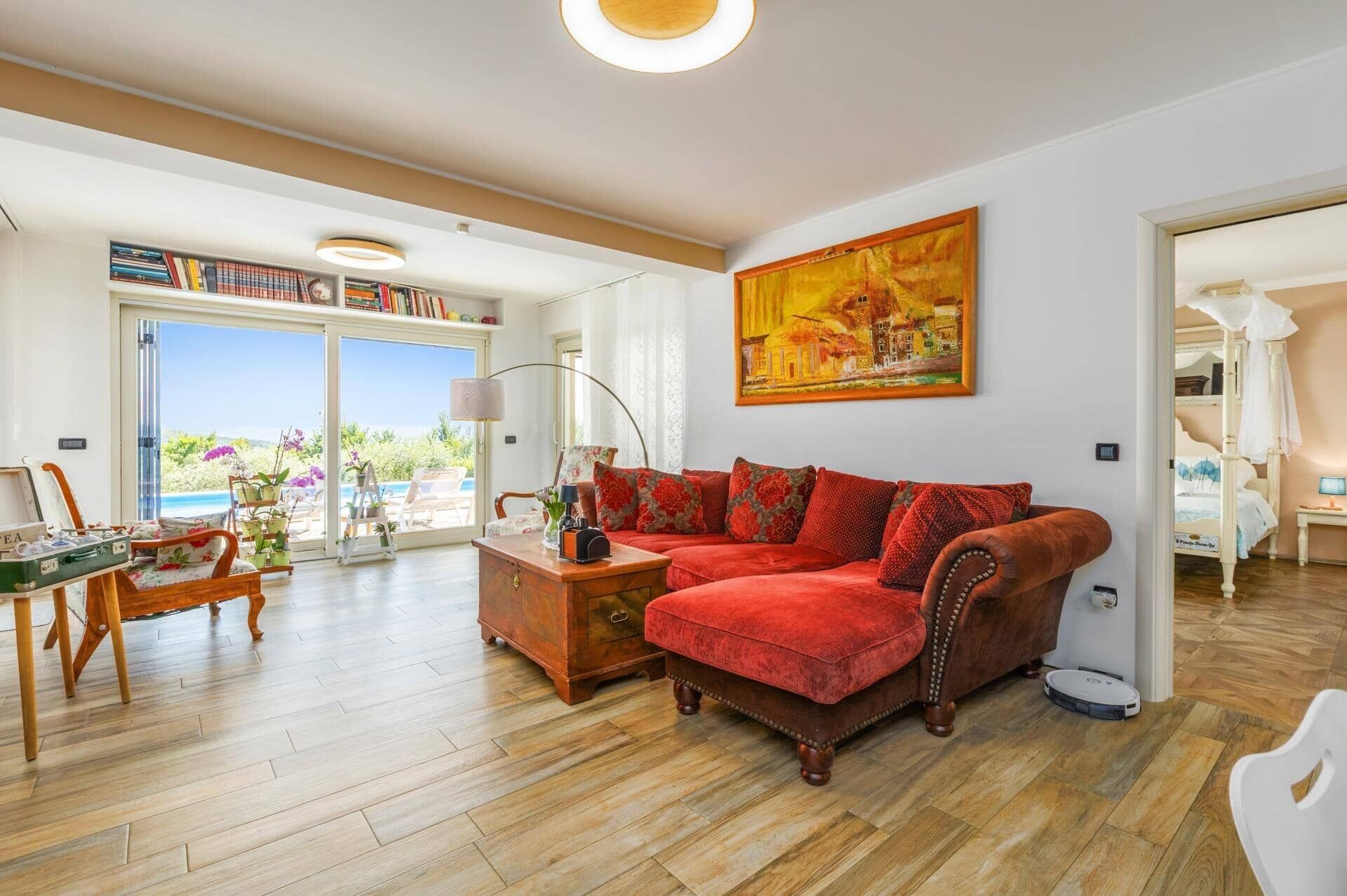 Un salon spacieux avec un canapé rouge, un parquet en bois, un tableau accroché au mur et une vue à travers la porte vitrée.