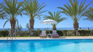 A pool area with two lounge chairs and a white umbrella, surrounded by palm trees and a view of the sea.