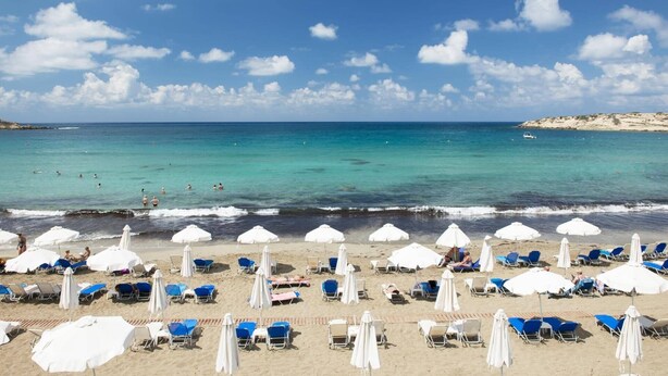 A beach with rows of sun loungers and umbrellas, a clear blue sea, and a sandy shore.