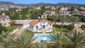 A white villa with a swimming pool, surrounded by palm trees and a view of a residential area with houses and mountains in the background.
