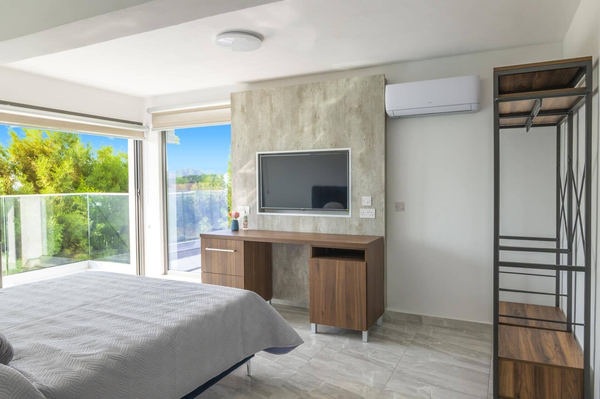 A modern bedroom with a bed, a TV mounted on a concrete wall, a wooden desk, and a balcony with a view of greenery.