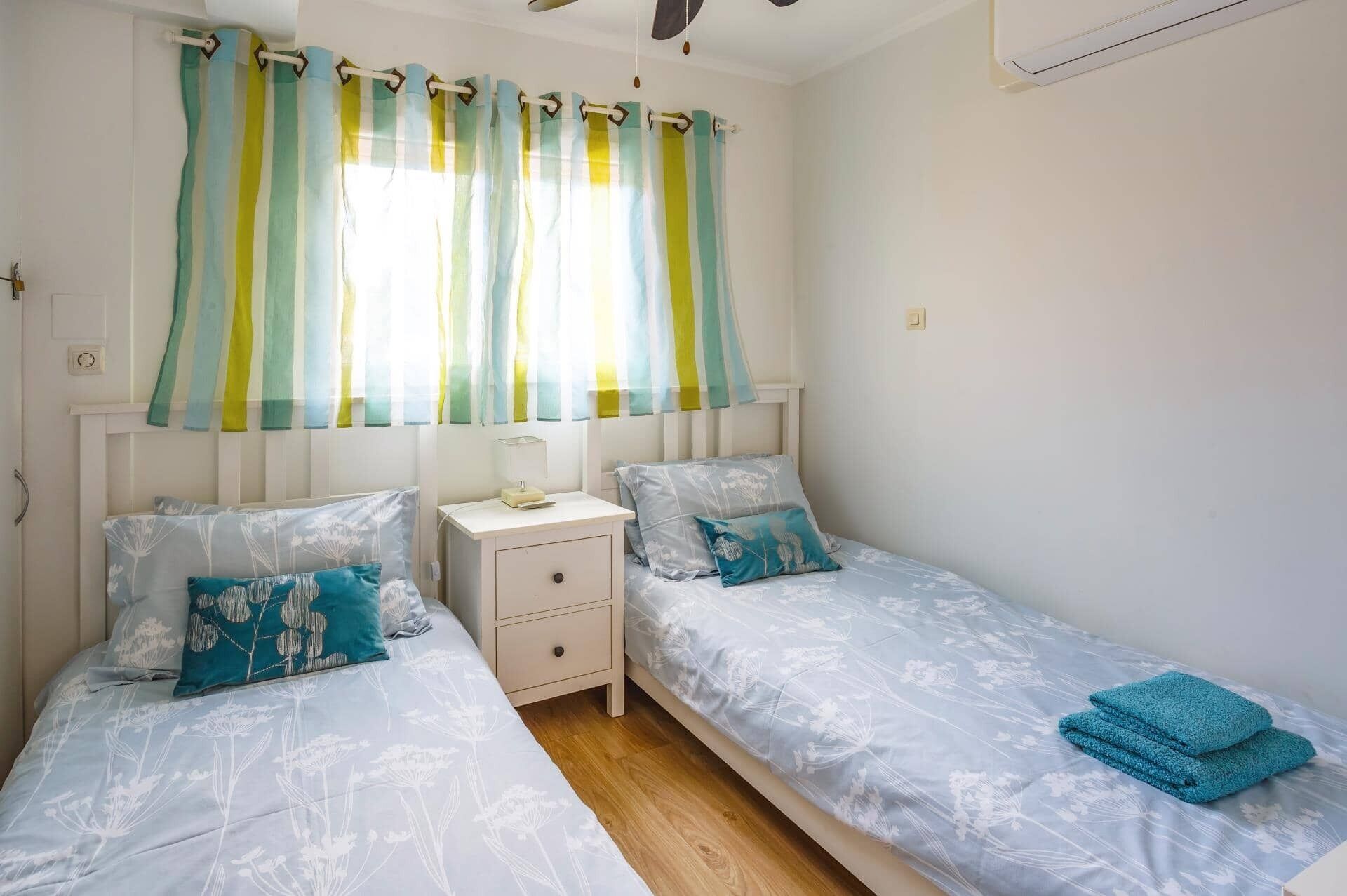 A bedroom with two single beds, a nightstand, a bedside table, and a window with striped curtains.