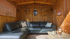 Interno di una baita in legno con un divano in pelle nera, un tavolino da caffè intrecciato con bicchieri e una bottiglia, e pareti in legno con diversi cartelli.