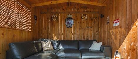 A wooden cabin interior with a black leather sofa, a woven coffee table with glasses and a bottle, and wooden walls with various signs.