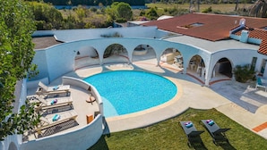 A swimming pool with a curved design, surrounded by lounge chairs and a covered patio area. - Villa Les Arches by Villa Plus (Villedaigne)