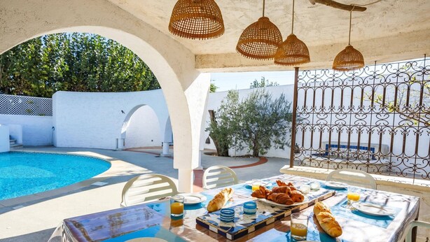 A poolside dining area with a table set for breakfast, including bread and juice, and a view of the pool and greenery. - Villa Les Arches by Villa Plus (Villedaigne)
