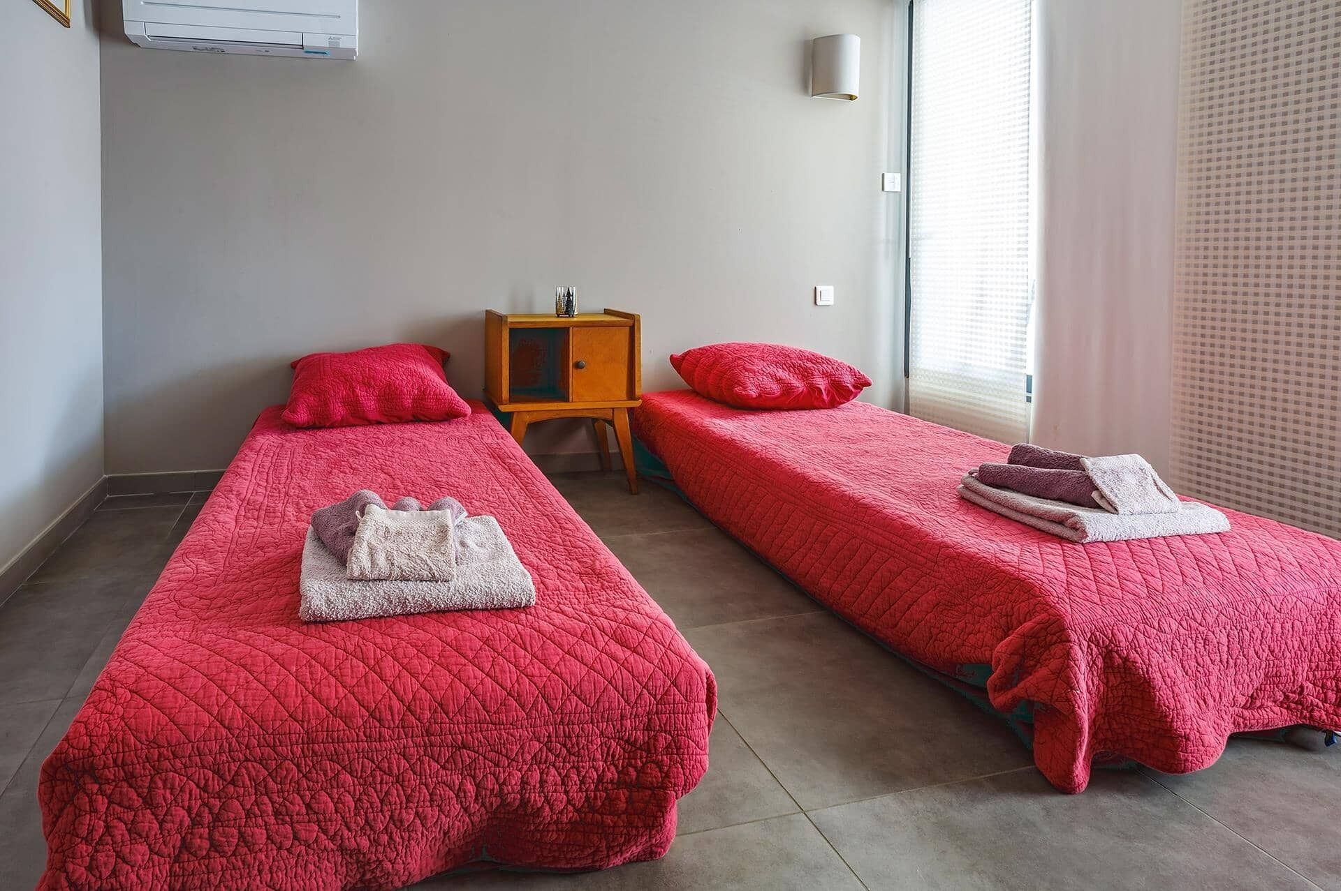 Due letti singoli con lenzuola rosse, un comodino in legno e una lampada a muro.