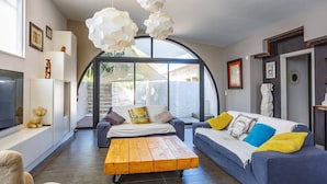 A modern living room with a large window, a wooden coffee table, two sofas with colorful cushions, and a unique ceiling light fixture. - Villa Saffron by Villa Plus (Villedaigne)