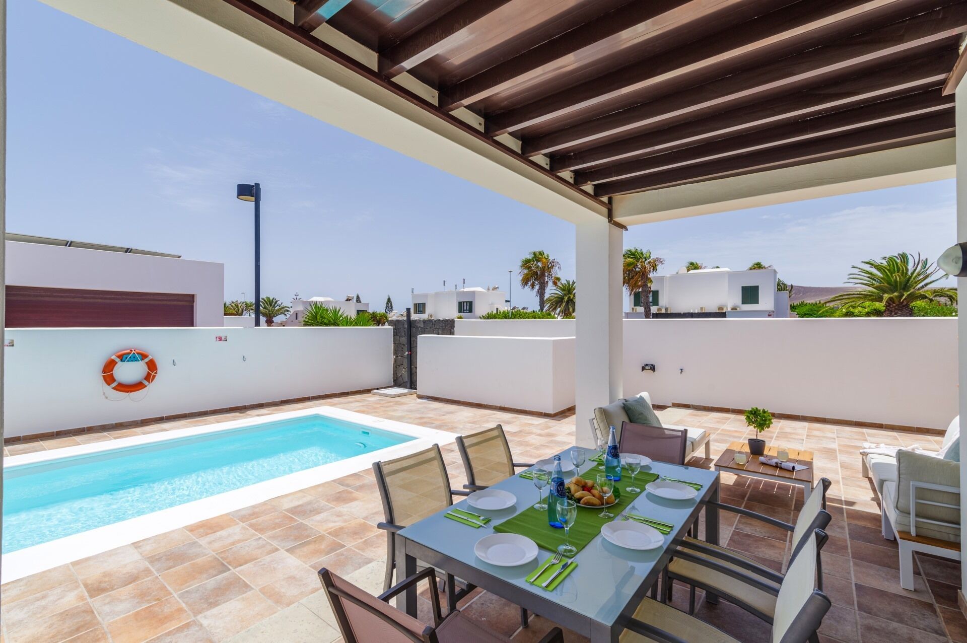 Área de piscina en la azotea con una mesa de comedor preparada para cuatro personas, rodeada de sillas y vistas al mar y edificios cercanos.