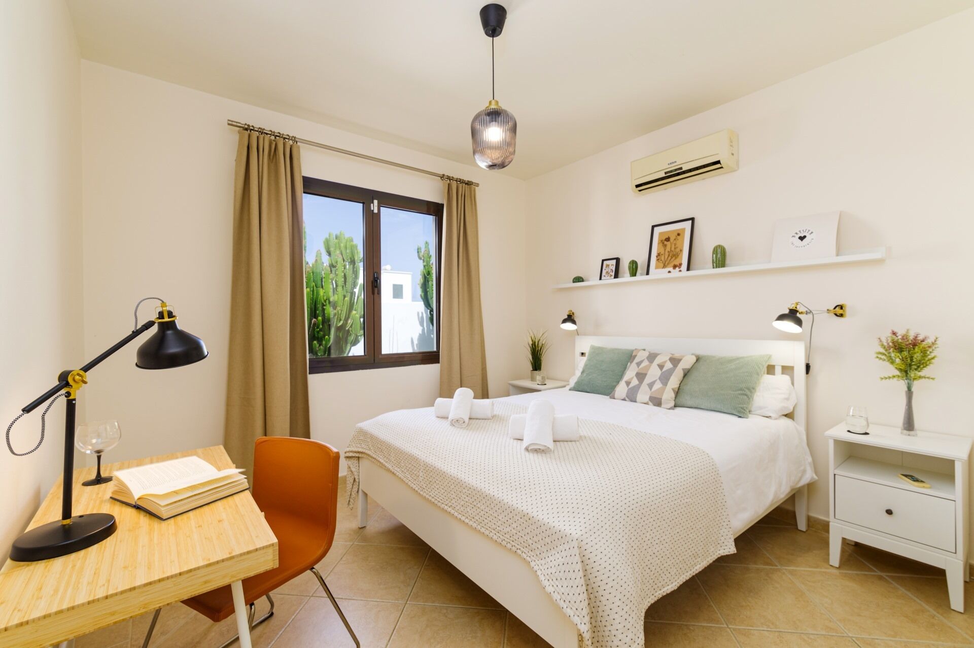 A bedroom with a bed, a desk with a lamp, a chair, a bedside table with a lamp, and a window with curtains.