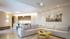 A modern living room with a sectional sofa, a small round table with chairs, a kitchen area in the background, and a large abstract painting on the wall. - Villa Lavender Bloom by Villa Plus (Lefkada)