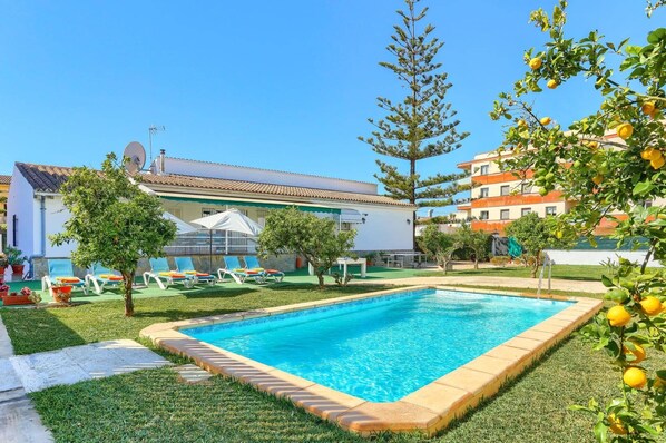 Une piscine entourée d’une pelouse bien entretenue, de quelques arbres et d’un bâtiment doté d’un toit recouvert de tuiles.