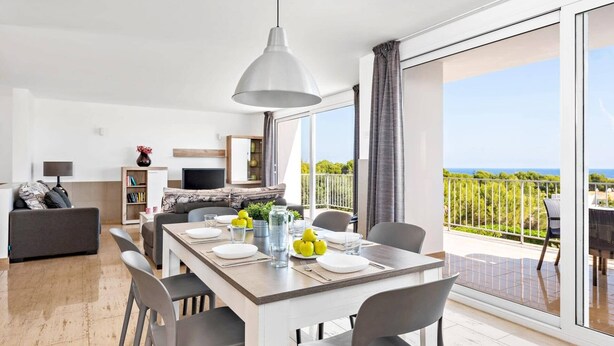A modern living room with a dining area, large sliding glass doors to a balcony, and a view of the sea. - Villa Grace by Villa Plus (Es Mercadal)