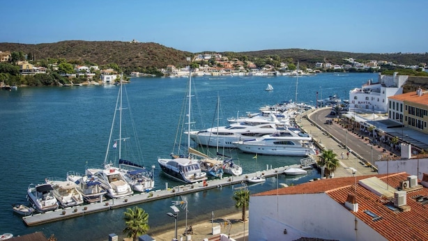 Un port de plaisance où sont amarrés de nombreux yachts, une ville côtière en arrière-plan et un ciel dégagé.