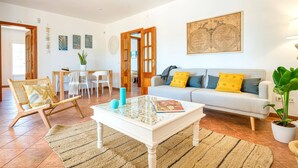 Un salon comprenant un canapé, une table basse et des fauteuils. On y trouve également un coin repas avec une table et des chaises. La pièce est dotée d’une grande fenêtre et d’une porte donnant sur l’extérieur.