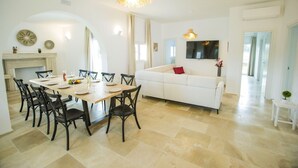 A dining area with a wooden table and chairs, a white sofa, and a TV mounted on the wall. - Villa Gabriella by Villa Plus (Carovigno)