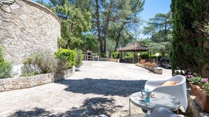 Un bâtiment en pierre doté d’un espace extérieur abrité avec des sièges, comprenant des chaises blanches et une table, le tout entouré de verdure et d’arbres.