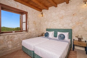 Quarto com parede de pedra, teto de madeira, uma cama grande com cabeceira verde, duas mesas de cabeceira com luminárias e uma janela com vista para o verde.
