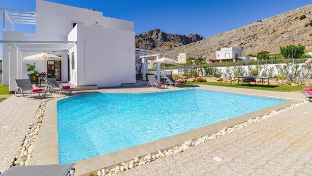 A swimming pool with lounge chairs and umbrellas, surrounded by a white building and a mountainous landscape. - Villa Pandora Lindos by Villa Plus (Rhodes)