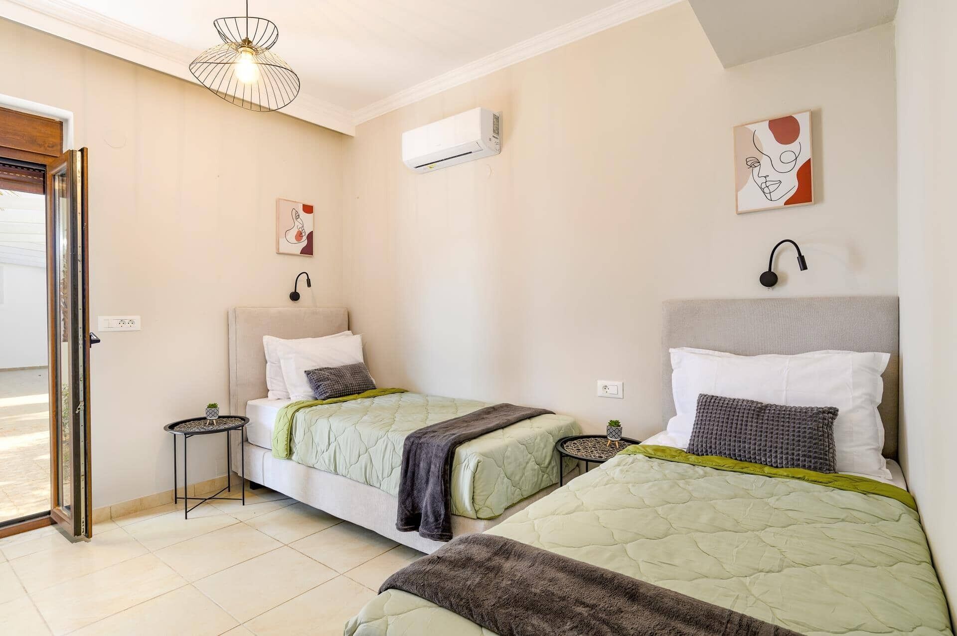 A bedroom with two beds, a small table, a wall-mounted air conditioner, and a ceiling light fixture.