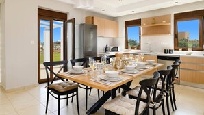 A modern kitchen with a dining area set for six, featuring a large island, stainless steel appliances, and a view of the outdoors. - Villa Pandora Lindos by Villa Plus (Rhodes)