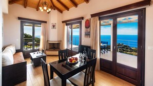 A spacious living area with a fireplace, a dining table set for two, and a sofa. Large sliding glass doors lead to a balcony with a view of the sea. - Villa Drimonas Erato by Villa Plus (Lefkada)