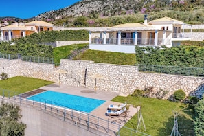 Zona de piscina con pared de piedra, sillas de descanso y vistas a casas en la ladera.
