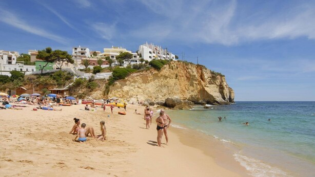 A beach with people sunbathing and swimming, a rocky cliff in the background, and buildings on the hillside. - Villa Curva do Monte by Villa Plus (Praia do Carvoeiro)