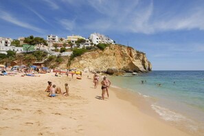 A beach with people sunbathing and swimming, a cliff with buildings, and clear skies.