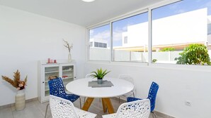 A dining area with a round table, blue and white chairs, a white cabinet with glass doors, and a view of buildings and greenery through large windows. - Villa Naya by Villa Plus (Playa Blanca)