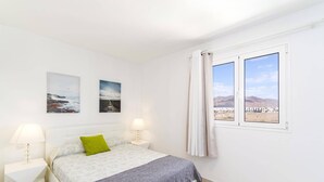 A bedroom with a bed, two bedside lamps, a window with a view, and two wall-mounted pictures. - Villa Naya by Villa Plus (Playa Blanca)