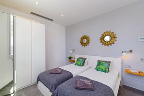 Two single beds with grey bedding and decorative pillows, a wardrobe, and a bedside table with a lamp.