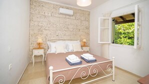A bedroom with a stone wall, a bed with white bedding, two bedside tables with lamps, and a window with open shutters. - Villa Mistral Cottage by Villa Plus (Ionian Islands)