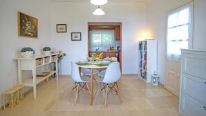 A dining area with a table set for four, white chairs, a white bookshelf, and a kitchen in the background. - Villa Mistral Cottage by Villa Plus (Ionian Islands)