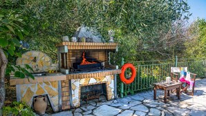 An outdoor barbecue area with a stone fireplace, a fire burning, and a green fence. - Villa Mistral Cottage by Villa Plus (Ionian Islands)