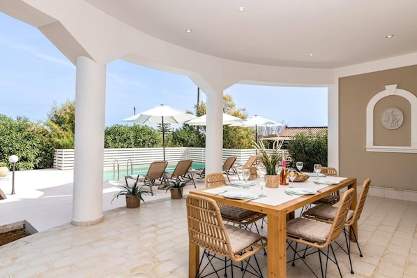 A covered outdoor dining area with a pool, lounge chairs, and umbrellas.