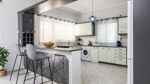 A modern kitchen with a marble island, bar stools, a washing machine, and a large window with sheer curtains.