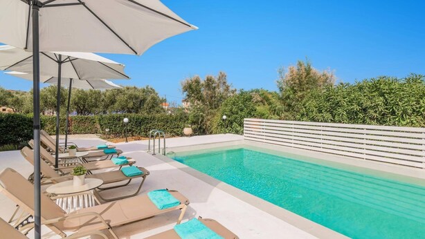 A swimming pool with lounge chairs and umbrellas, surrounded by greenery and a clear sky.