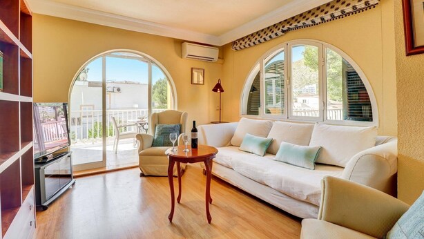 Una sala de estar con un sofá, una mesita y un televisor. Hay dos ventanas amplias con arcos y un equipo de aire acondicionado en la pared.