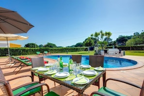 Un área de comedor junto a la piscina con una mesa dispuesta para cuatro personas, rodeada de sillas de mimbre y un gran paraguas.