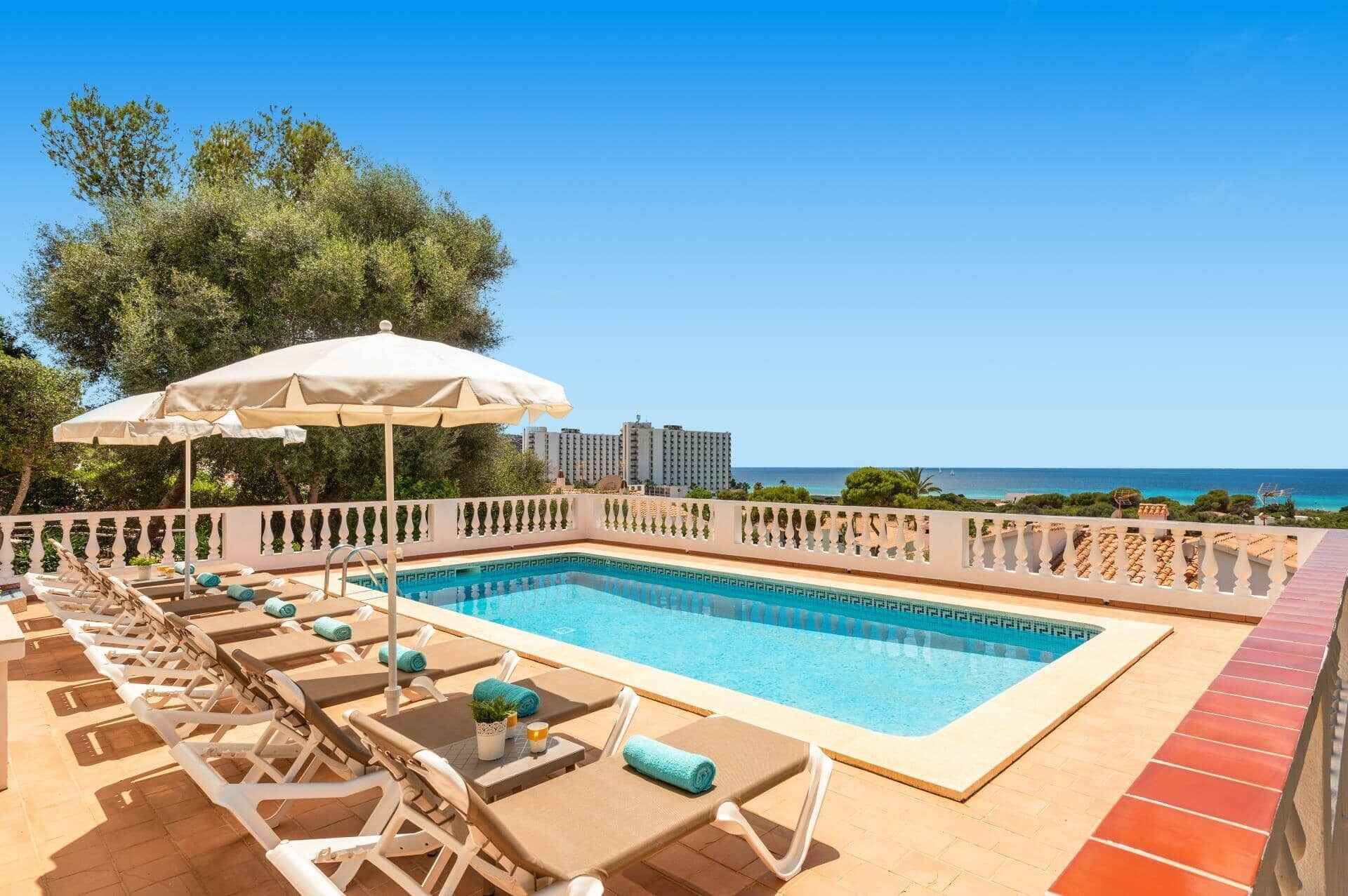 A poolside area with lounge chairs and a large umbrella, overlooking a clear blue sky and a distant view of the sea.