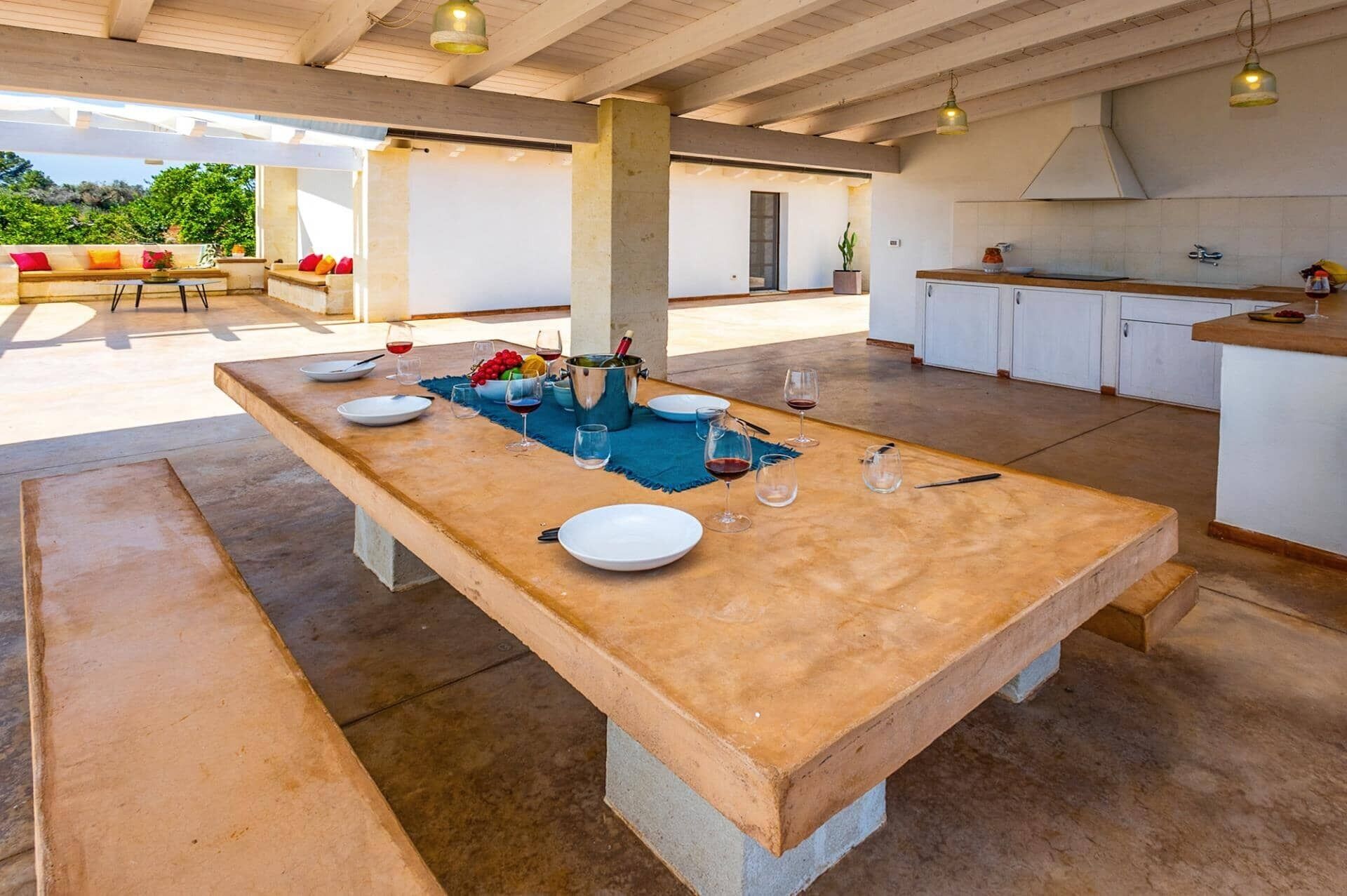 Un amplio espacio al aire libre con comedor, una mesa larga dispuesta para seis personas, rodeado de una cocina y una zona de estar con cojines.