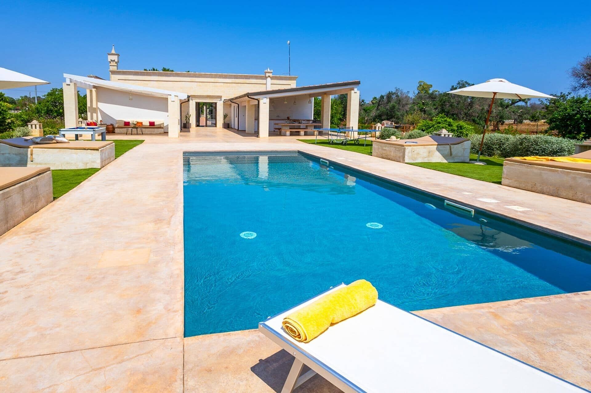Una piscina con una tumbona y una toalla, rodeada de un patio con mobiliario exterior y un edificio al fondo.