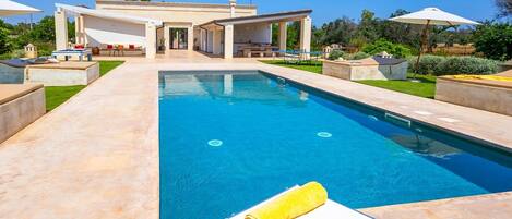 Una piscina con una tumbona y una toalla, rodeada de un patio con mobiliario exterior y un edificio al fondo.