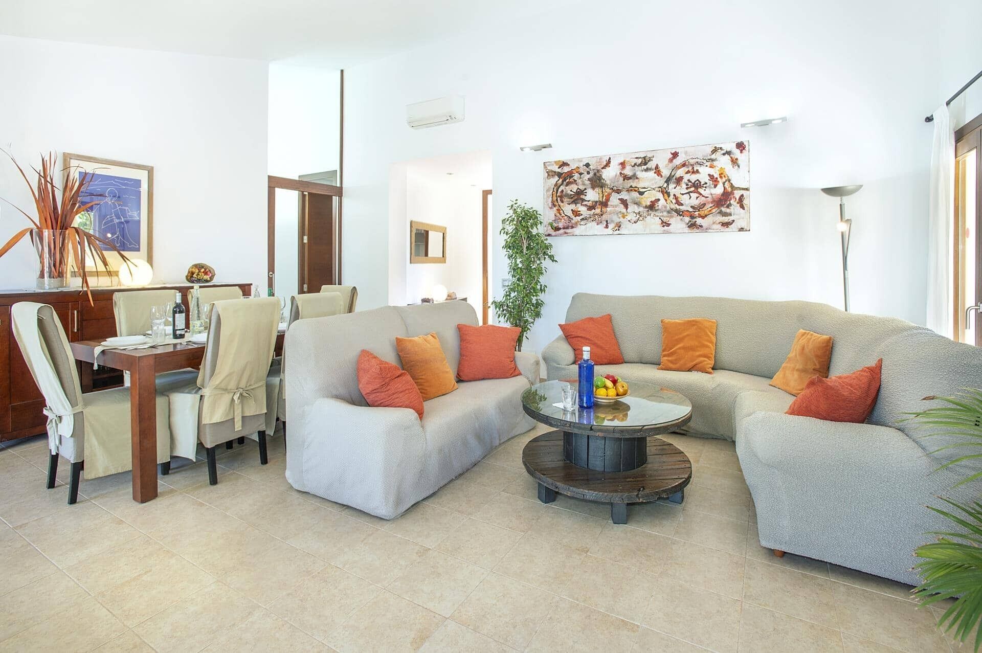 Un salon avec un canapé gris, des coussins orange, une table basse ronde et un coin repas comprenant des chaises et une table.