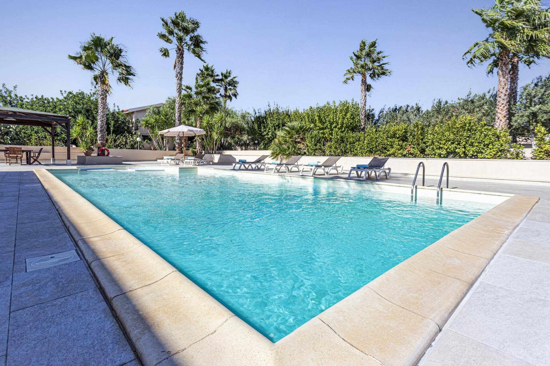 A rectangular pool with clear blue water, surrounded by lounge chairs and palm trees.